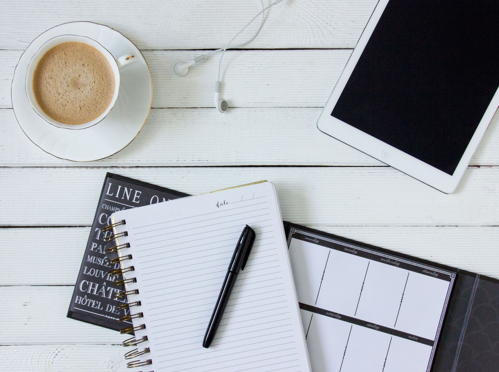 Flat lay of a workspace with notebook, tablet, coffee, and earphones on a white desk.
