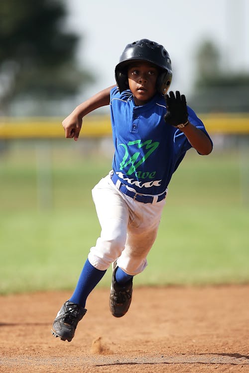 Gratis Boy In Blue And White Baseball Jersey Running On Brown Soil Field Selama Siang Hari Foto Stok
