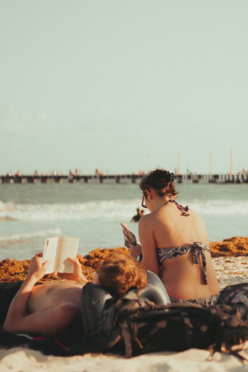 Free Two adults reading on a sandy beach with a sea view and distant pier. Stock Photo
