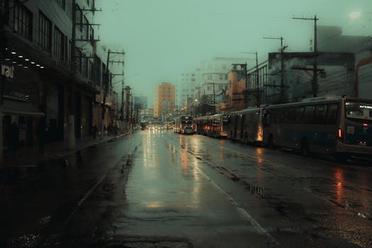 A moody, wet city street with buses lined up under dim lighting, reflecting rain-soaked pavement.