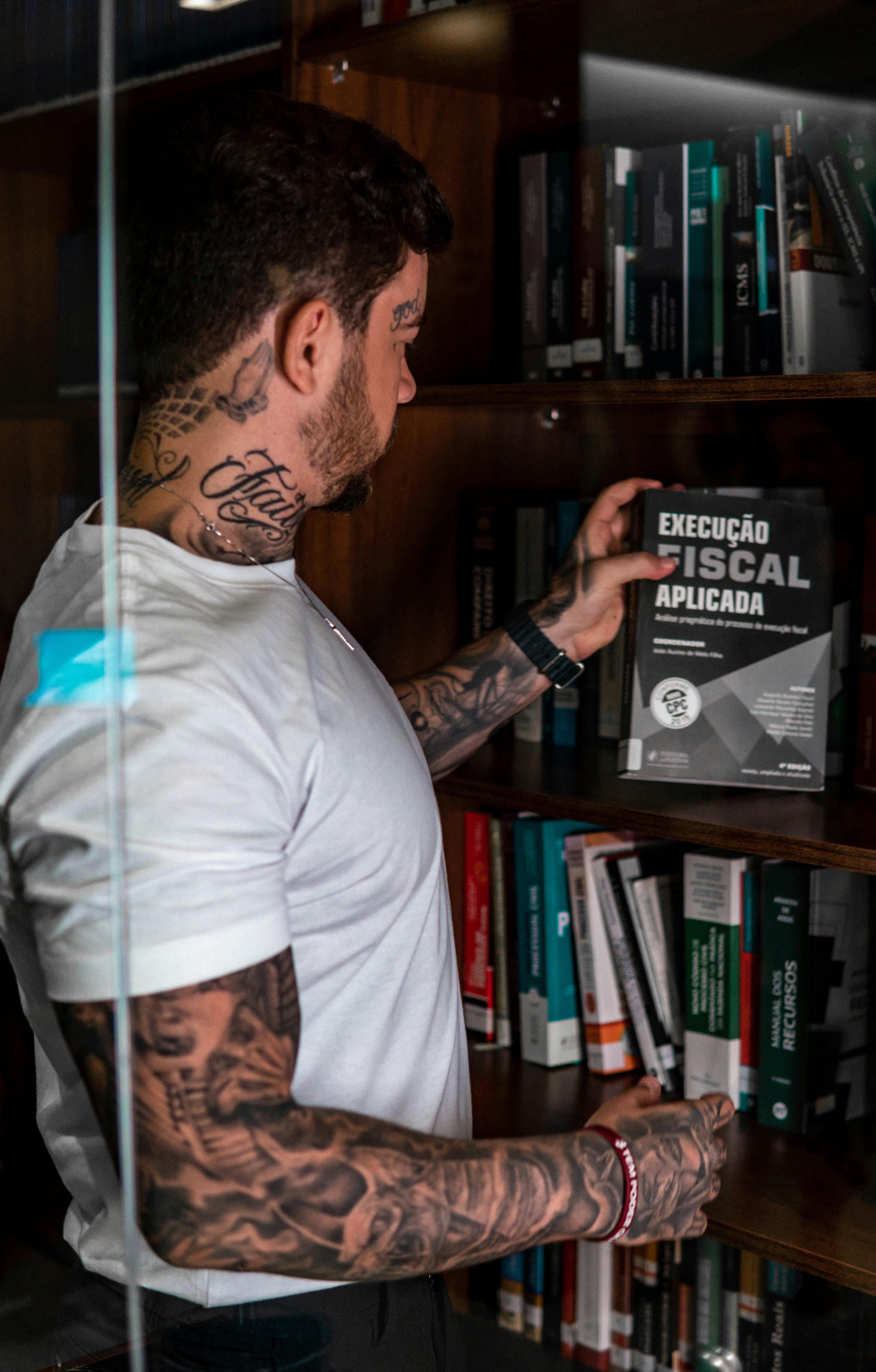 A tattooed man in a white t-shirt browsing a library bookshelf with focus on law books.