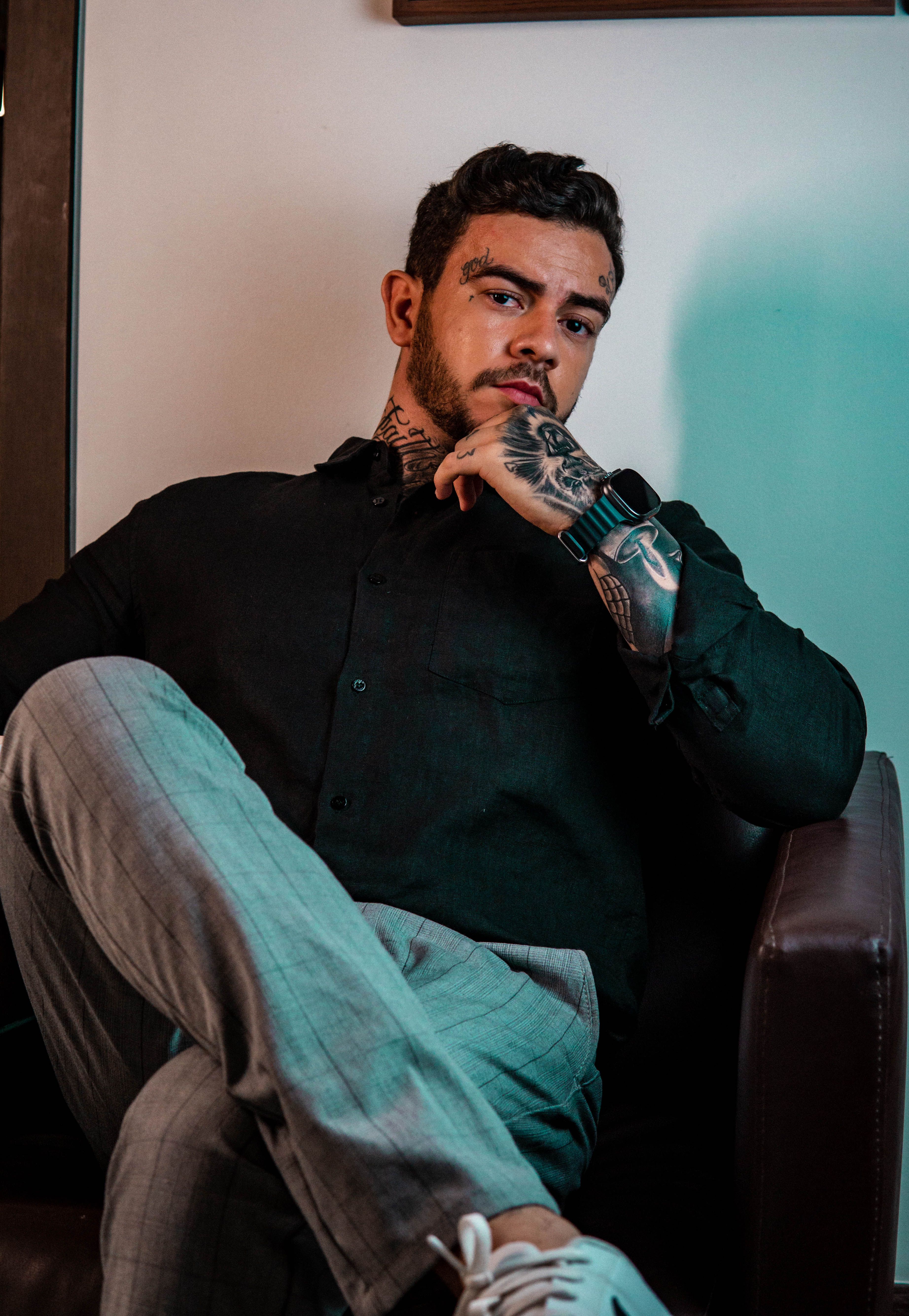 Elegant tattooed man in black shirt and gray pants posing in an armchair indoors.