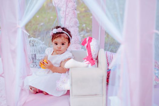Cute baby girl in a white dress with a floral headband, playing with toys in a pink canopy.