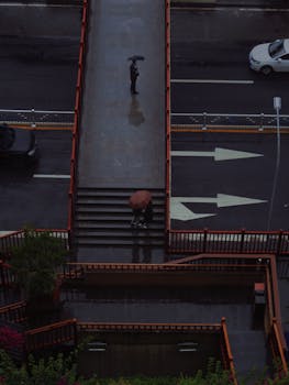 Aerial shot of people walking with umbrellas on a rainy urban road crossing a footbridge.
