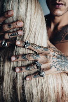 Close-up of tattooed hands with rings holding blonde hair, showcasing intricate tattoos and edgy style.