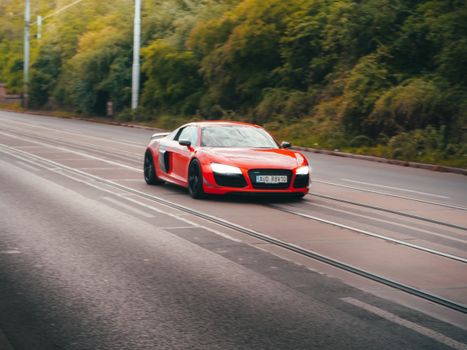 A sleek red sports car, Audi R8, speeding on a scenic road in Prague, Czechia.