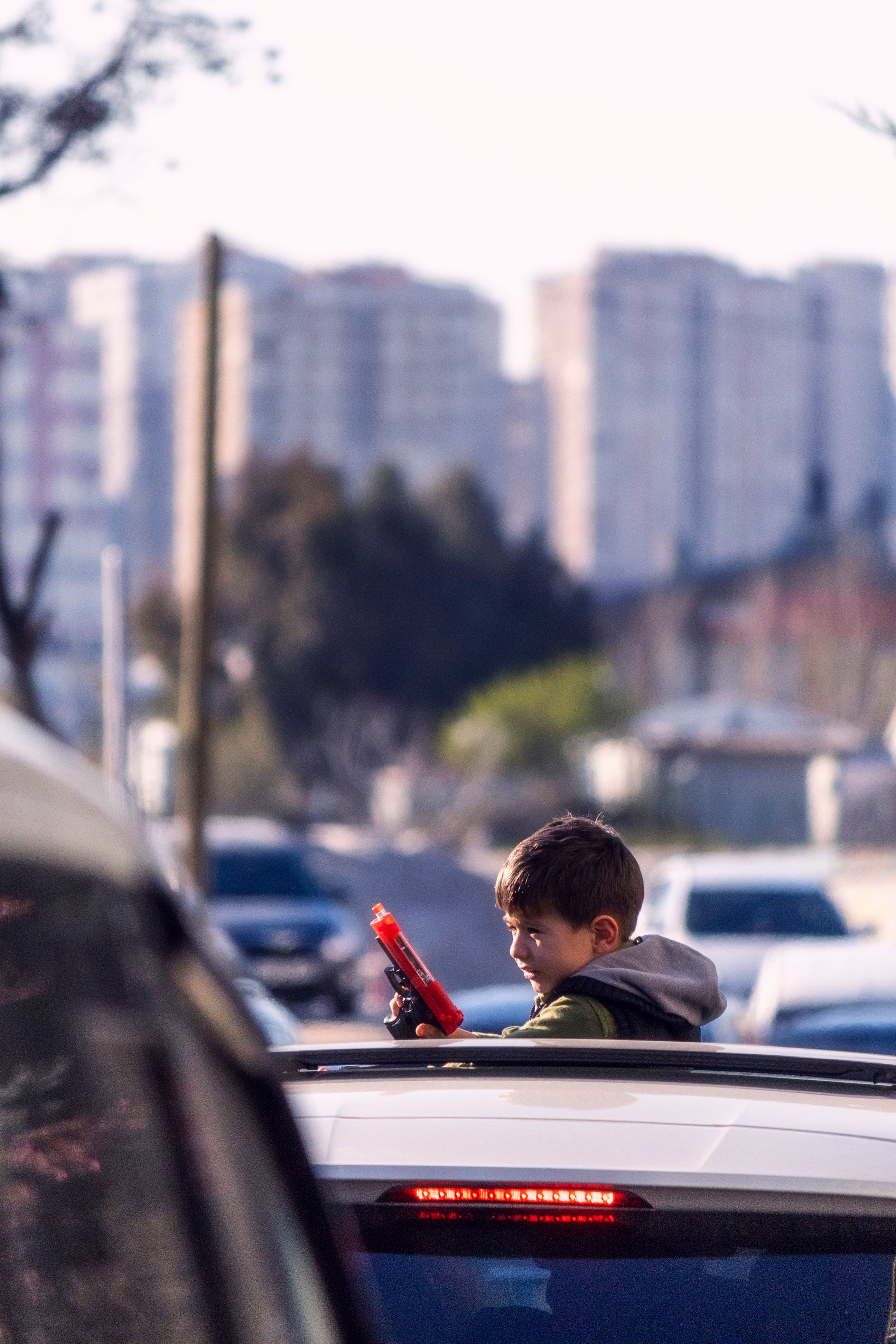 A young boy standing through a car sunroof holding a toy gun with a city skyline in the background.