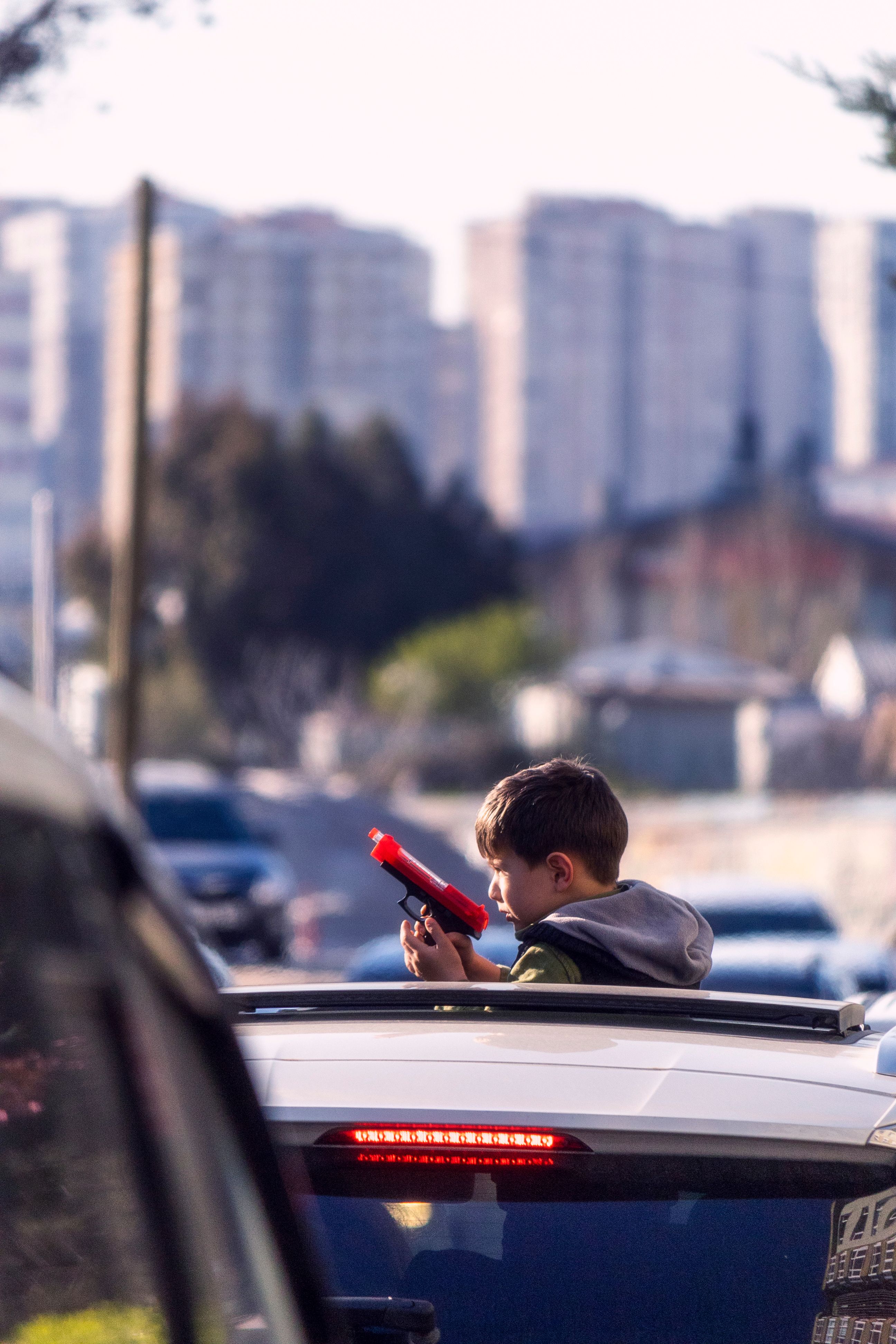 A young boy holds a toy gun while standing in a car on a busy urban street.