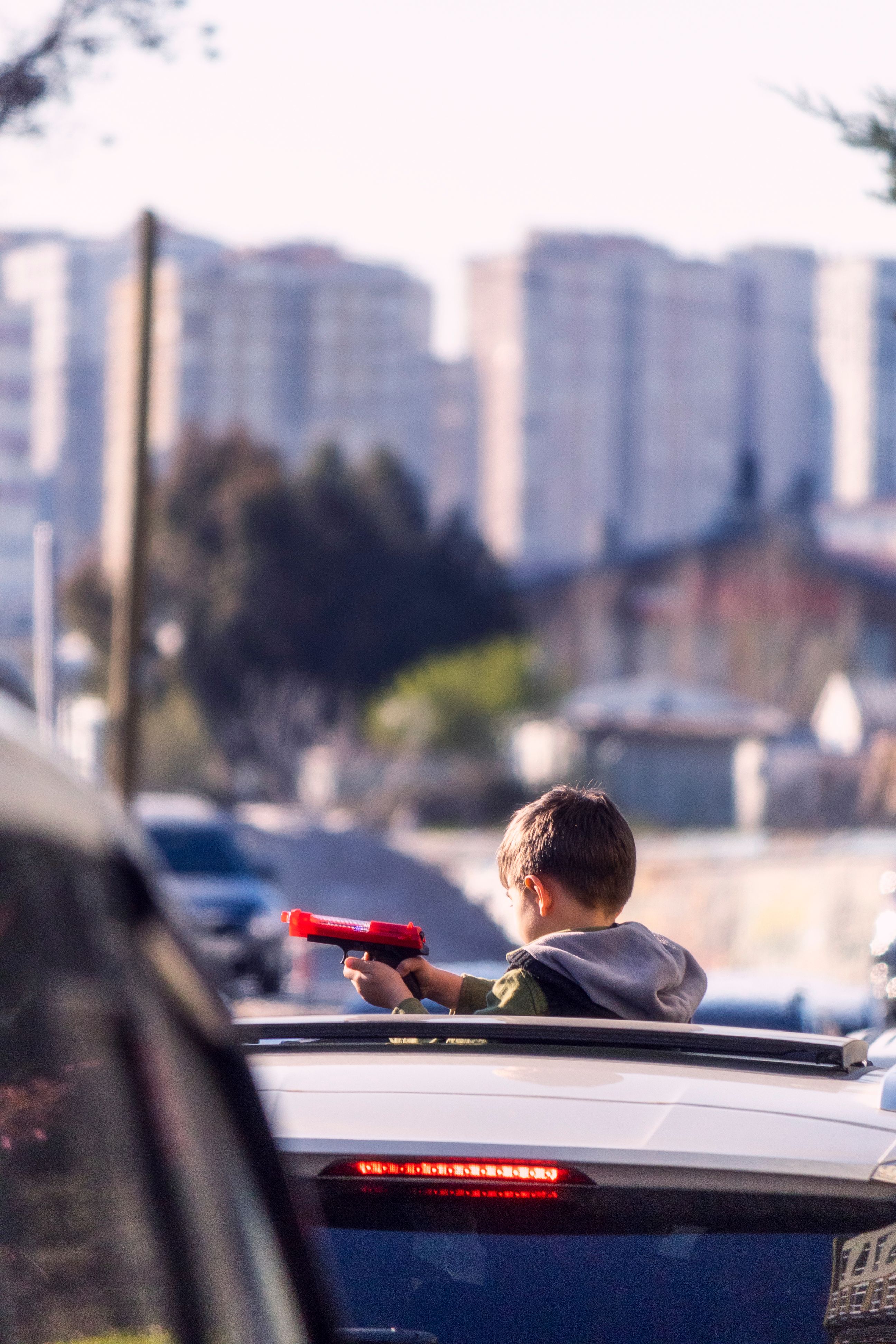 Young boy holding toy gun from car rooftop in city backdrop, sunny day.