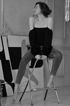Chic black and white portrait of a woman smiling while sitting on a chair in a casual outfit.