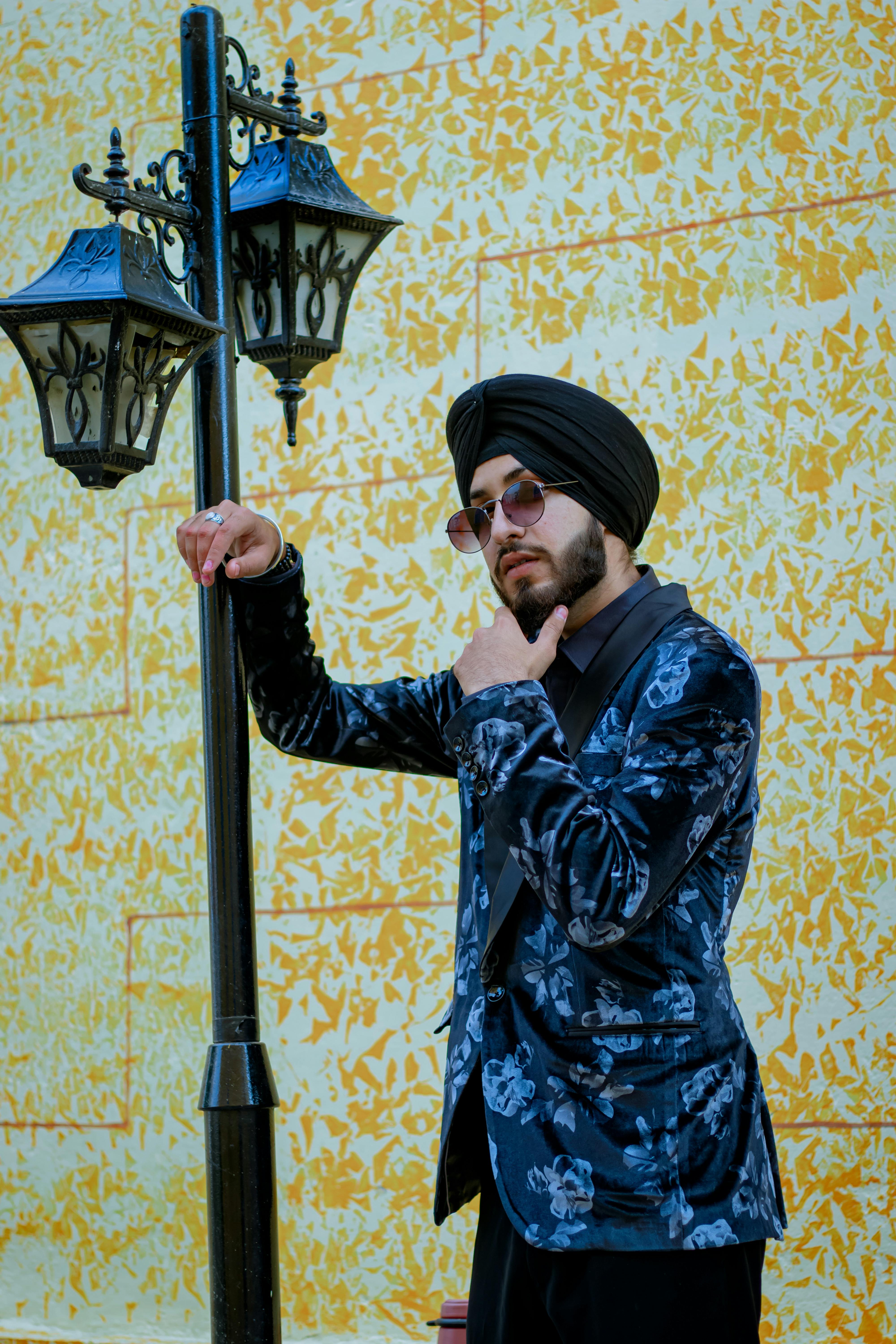 Portrait of a stylish young man in sunglasses and a turban, leaning against a lamppost outdoors.