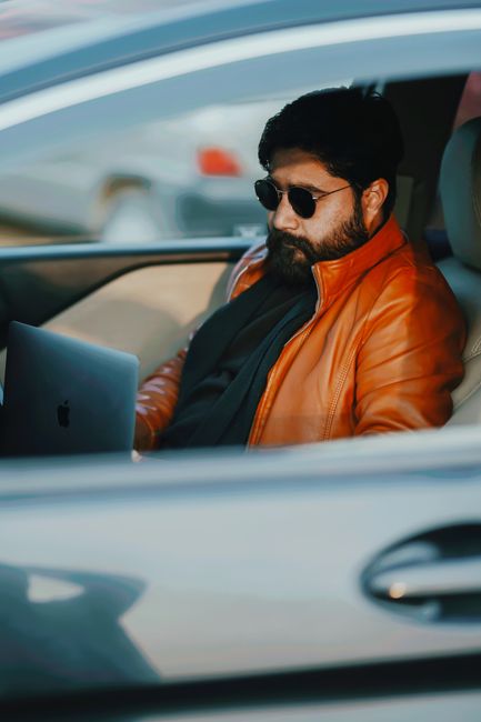 Man wearing sunglasses and leather jacket working on a laptop inside a car.