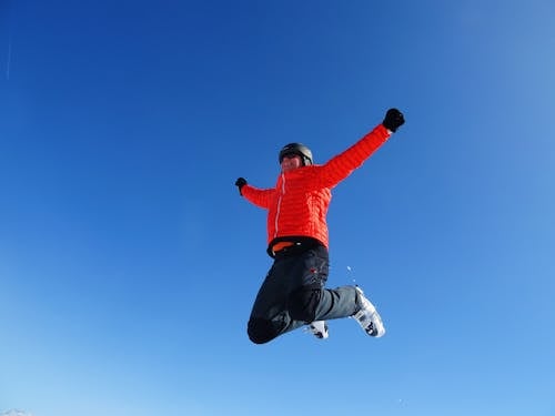 Man in Orange Zip Jacket and Black Pants Jumping Under Blue Sunny Sky