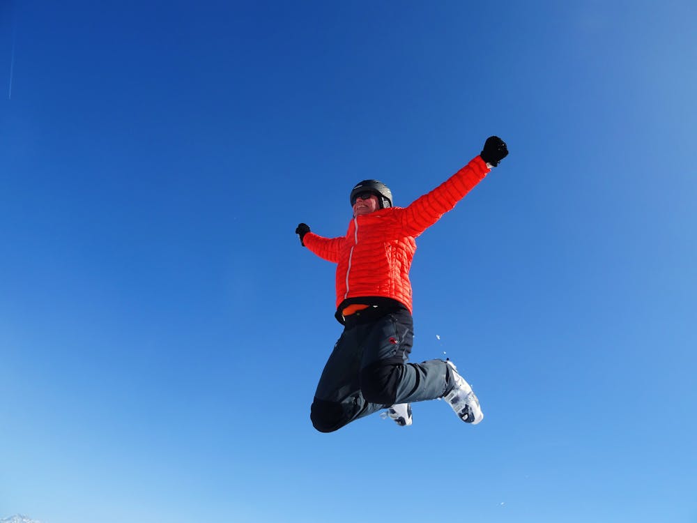 Free Man in Orange Zip Jacket and Black Pants Jumping Under Blue Sunny Sky Stock Photo