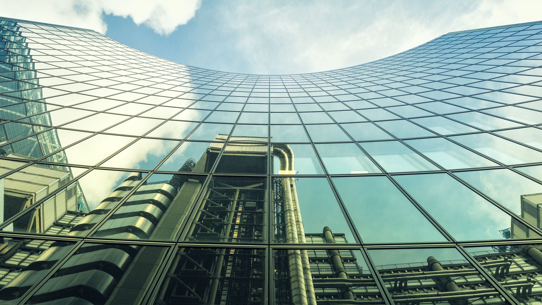 Low angle view of a modern glass building reflecting an industrial structure and sky.