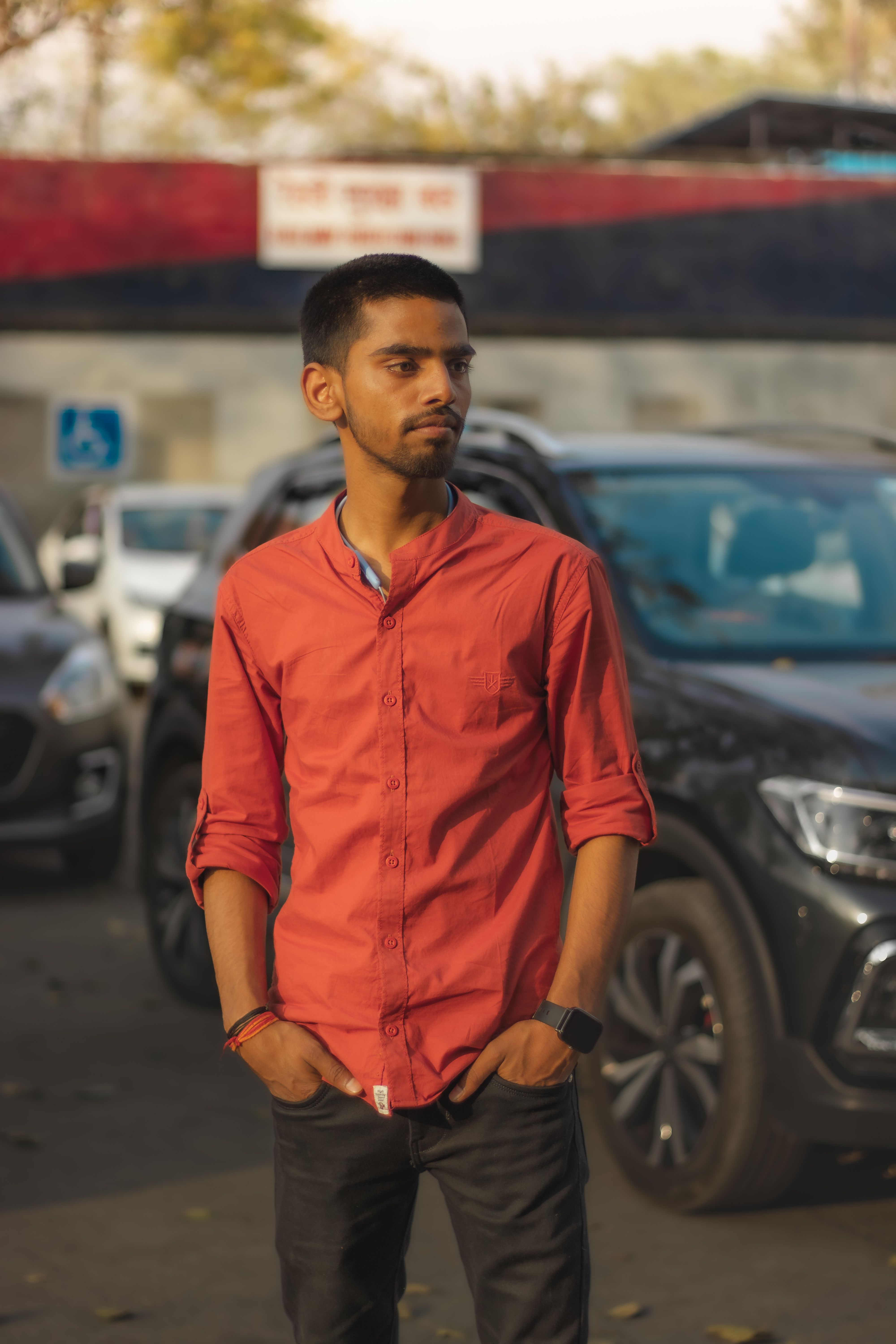 A young man in a red shirt poses in an urban area surrounded by cars, showcasing casual fashion.