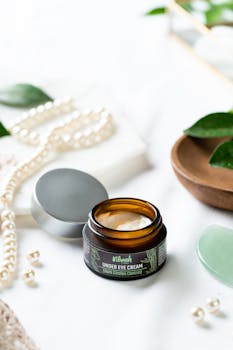 Elegantly styled under-eye cream jar with pearls and leaves on a white surface.