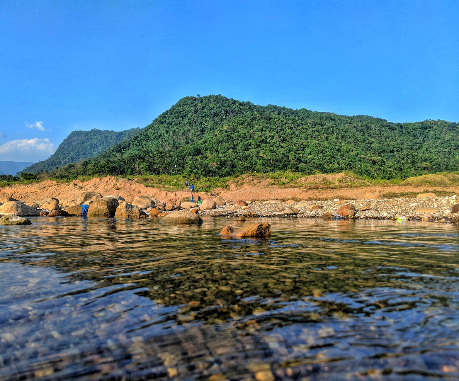 Explore the serene beauty of Sylhet District with lush hills and a clear river under a blue sky.