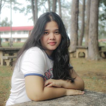 Portrait of a young woman sitting at a stone table in a park, surrounded by trees.
