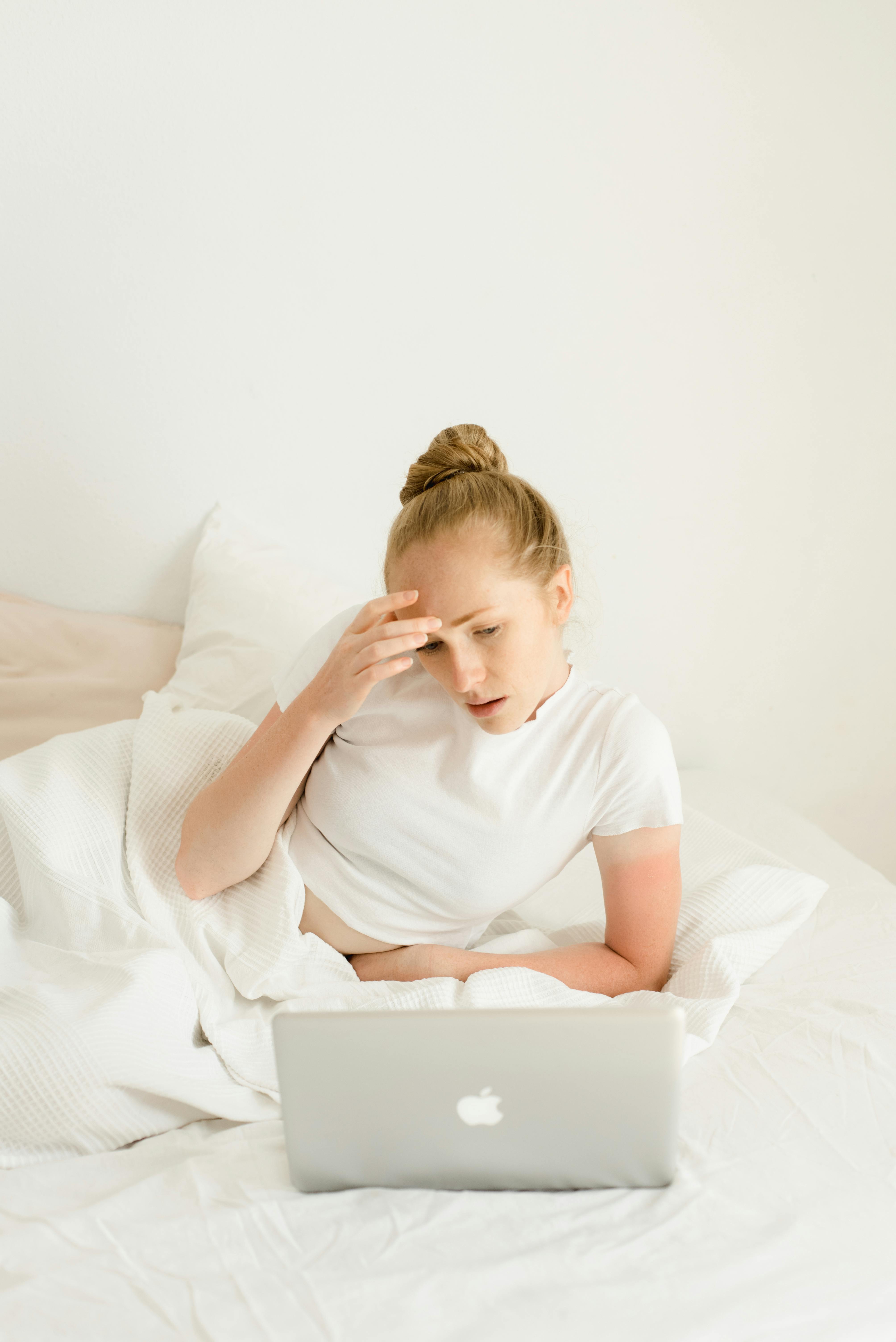 Young woman sitting in bed using a laptop, relaxed home environment with bright lighting.
