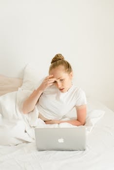 Young woman sitting in bed using a laptop, relaxed home environment with bright lighting.