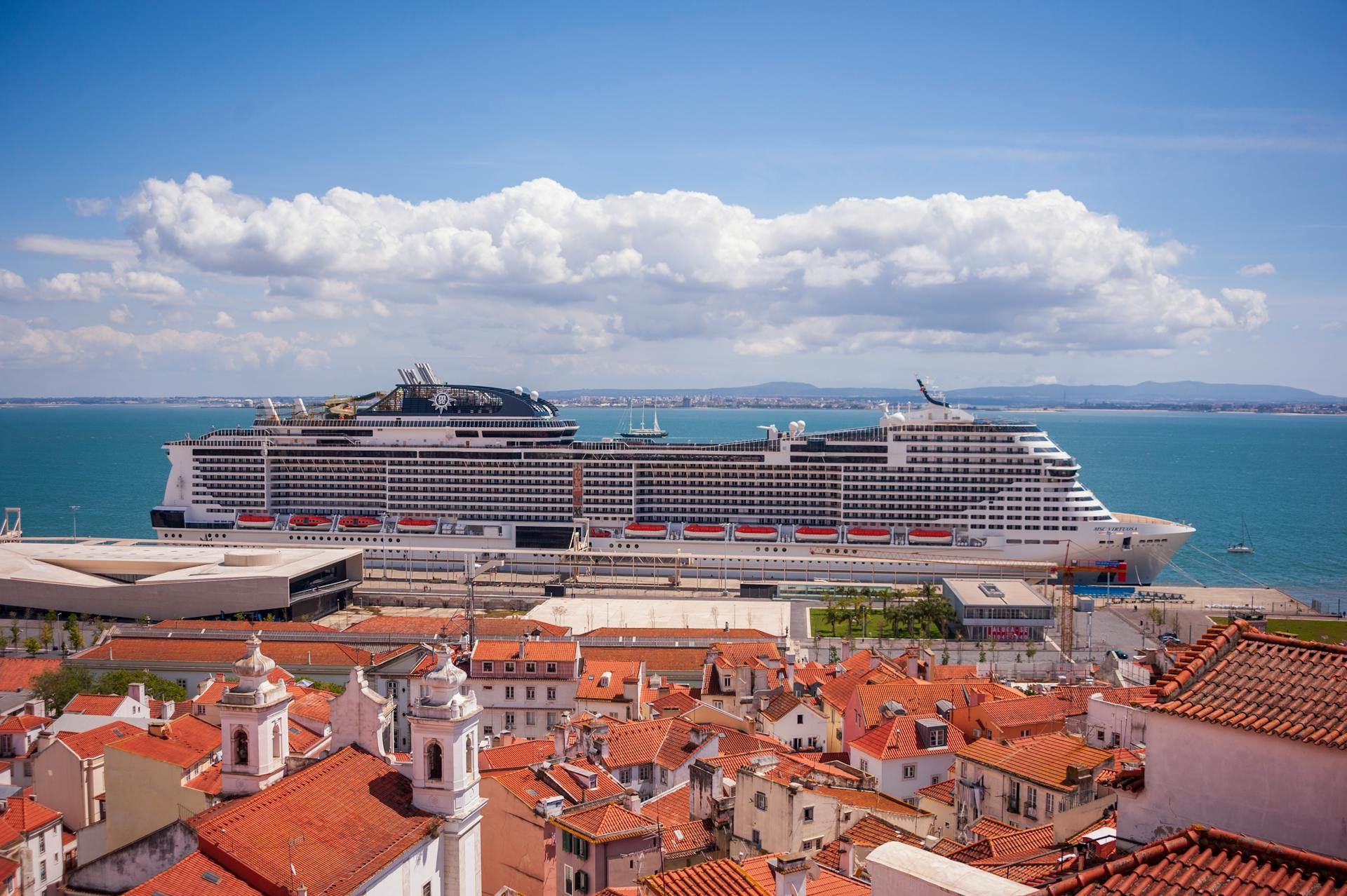 A large cruise ship docked at a scenic coastal town with red rooftops and a clear blue sky.
