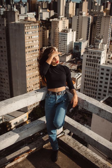 Woman in stylish black top and jeans poses on a rooftop overlooking a cityscape.