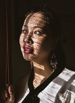 一位脸上有阴影图案的东亚妇女，正带着沉思的表情望向窗外