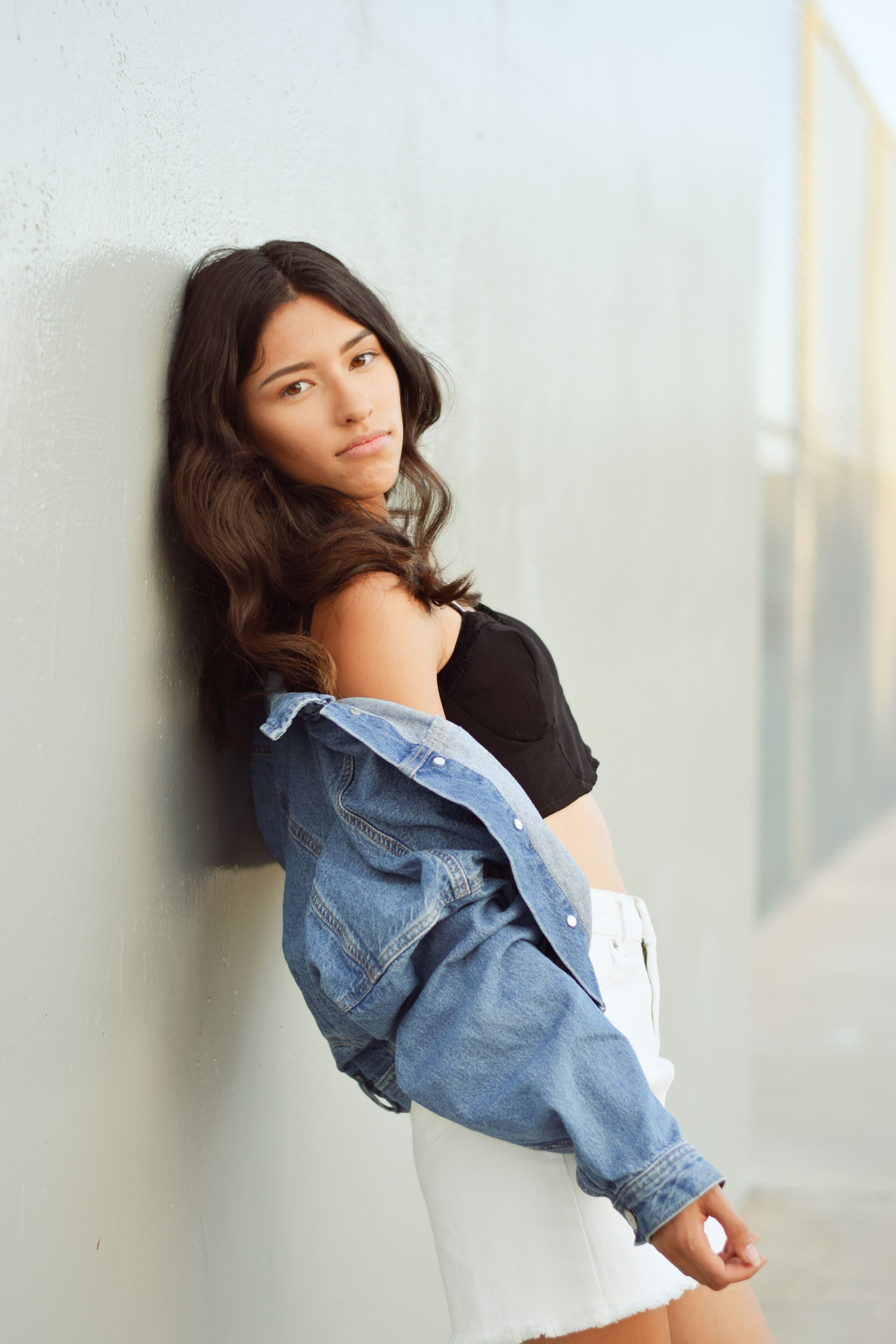 Stylish young woman leaning against a wall, wearing a denim jacket, black top, and white skirt in La Mirada, CA.