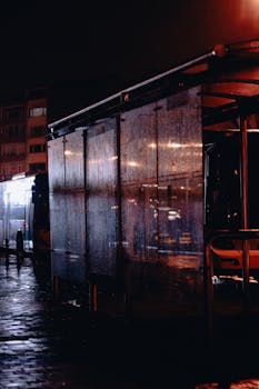 A rainy nighttime scene at an urban bus stop, capturing reflections and city lights.