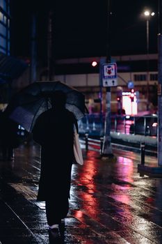 A person walks on a wet city sidewalk under an umbrella, reflecting street lights at night.