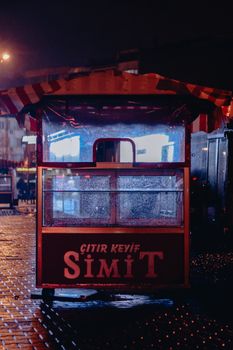 A vibrant simit cart with illuminated signs on a rainy city street at night, capturing urban nightlife.