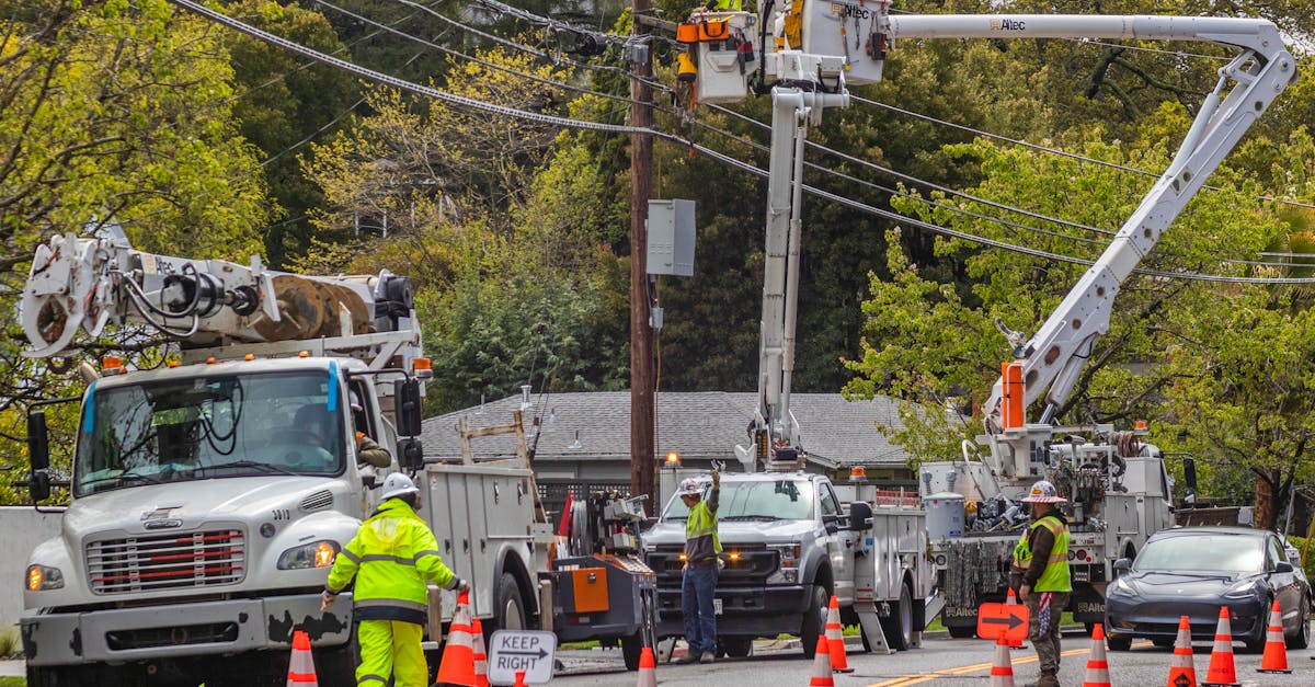 Photo by Robert So Utility workers in reflective gear repair power lines using bucket trucks and cones on a suburban street.