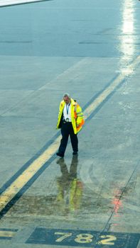 Ground crew member in reflective clothing walking on a rainy airport tarmac with wet surface reflection.