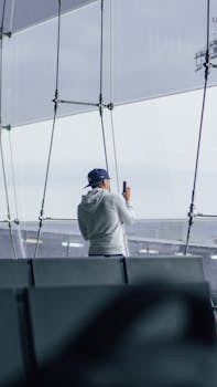 A man in a cap and hoodie using a smartphone to capture the view inside an airport terminal.
