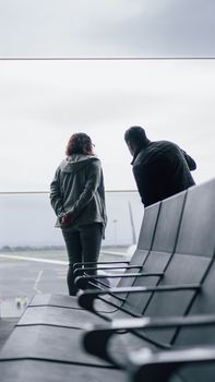 Two adults stand by airport window overlooking runway on an overcast day.