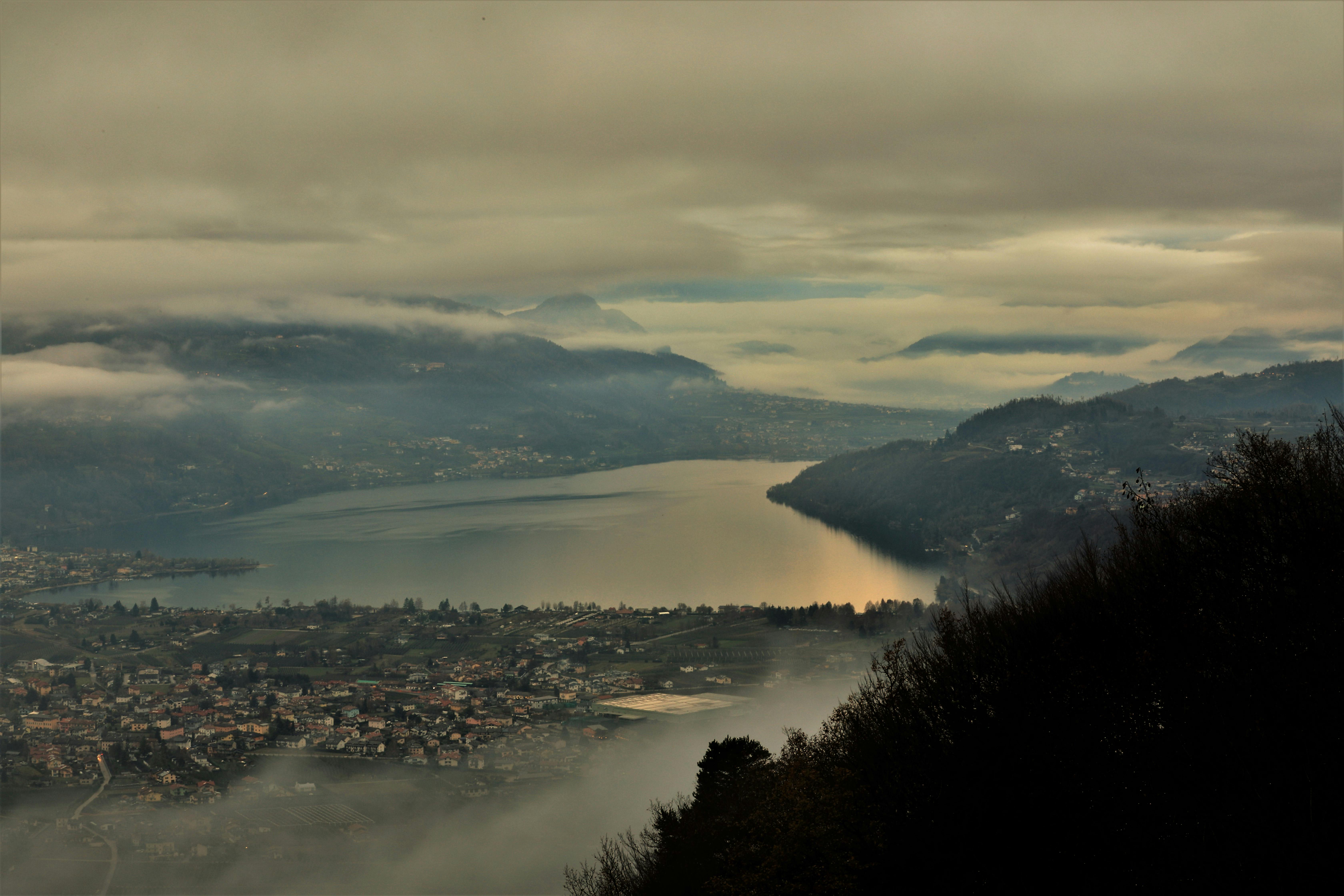 Aerial view of Trento, Italy, showcasing a misty landscape with lake, mountains, and villages under a cloudy sky.
