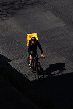 A food delivery cyclist with yellow backpack riding through urban streets, captured from a high angle.