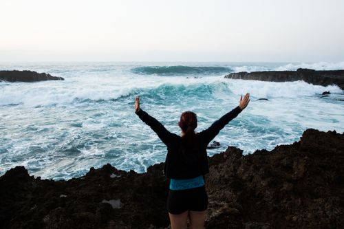Free A woman with arms raised stands on a rocky coast overlooking the ocean waves, enjoying the scenic view. Stock Photo