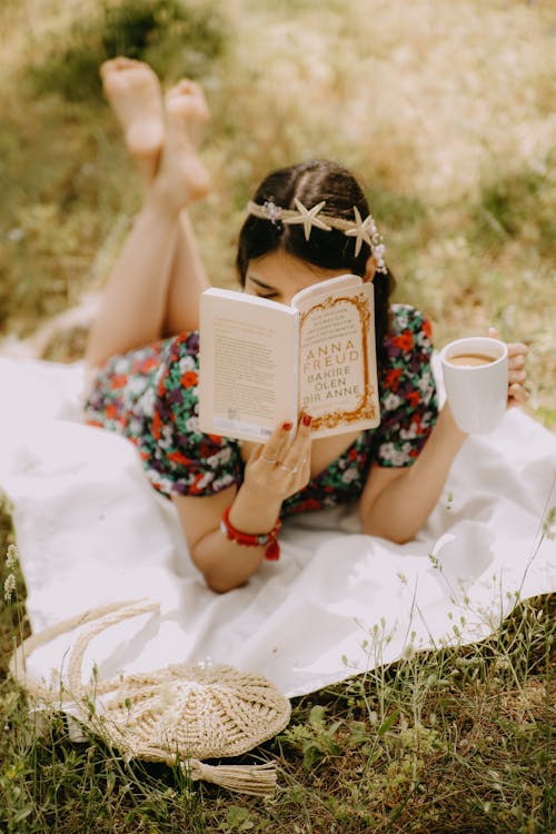 Young Woman Reading a Book and Drinking Coffee while Lying on a Picnic Blanket