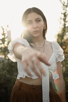 A young woman in sunlight reaching forward, wearing a white crop top outdoors.