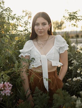 Fashionable woman in a white crop top posing outdoors surrounded by lush greenery and flowers.