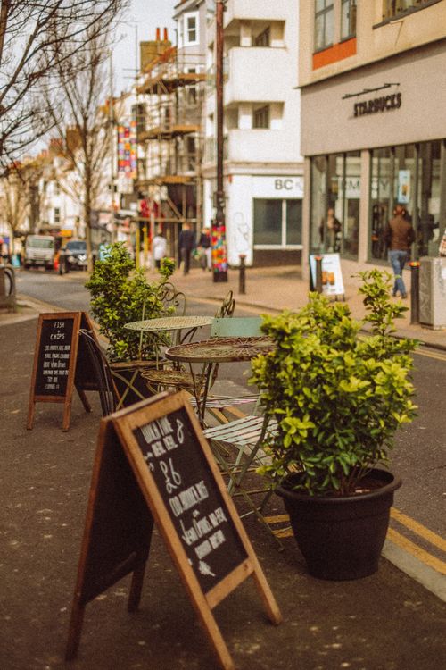 Free Inviting city street scene with café tables, potted plants, and storefronts. Ideal for lifestyle and travel themes. Stock Photo