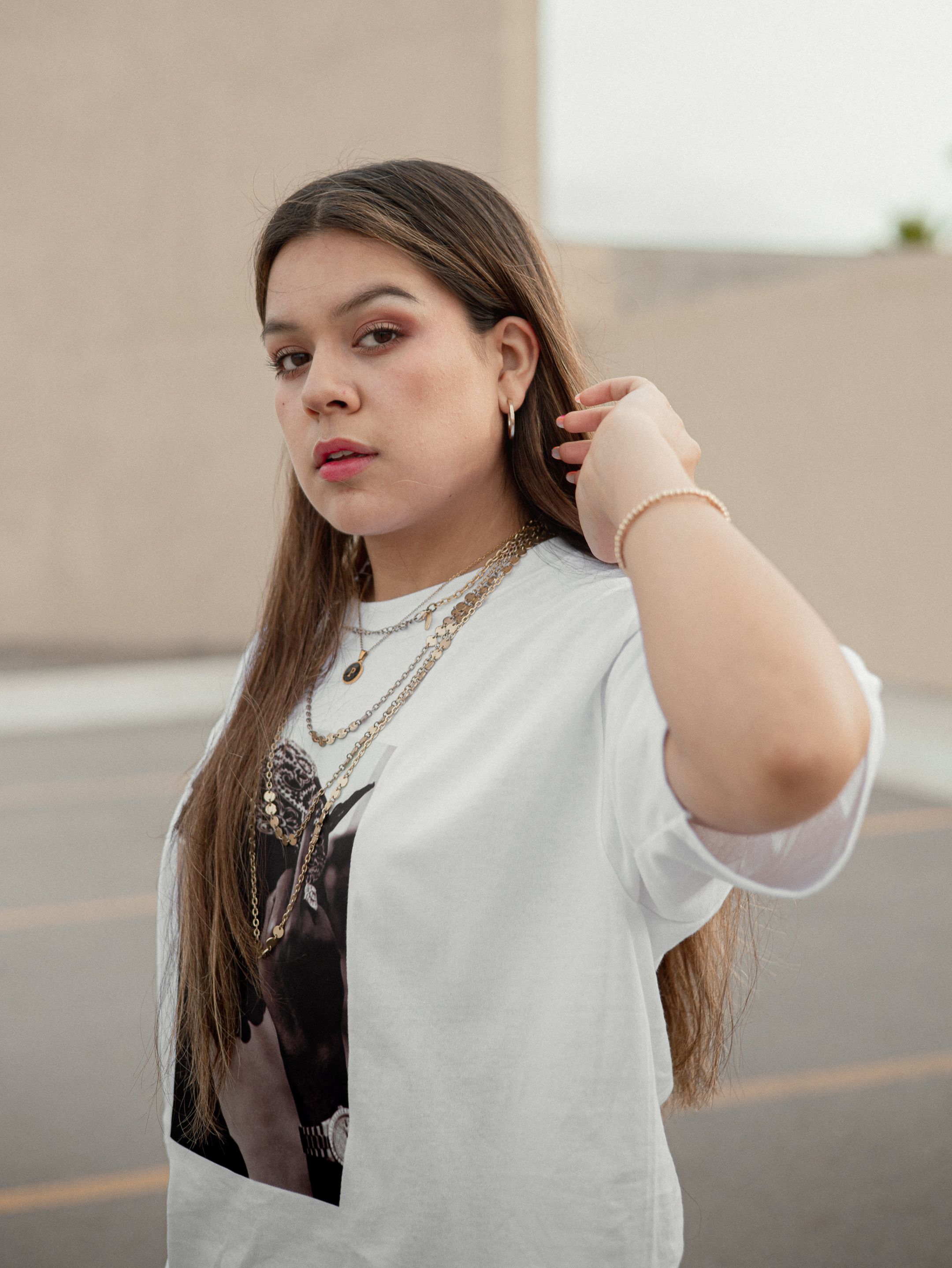 Young woman with long brown hair wearing a white t-shirt and jewelry in an urban outdoor scene.