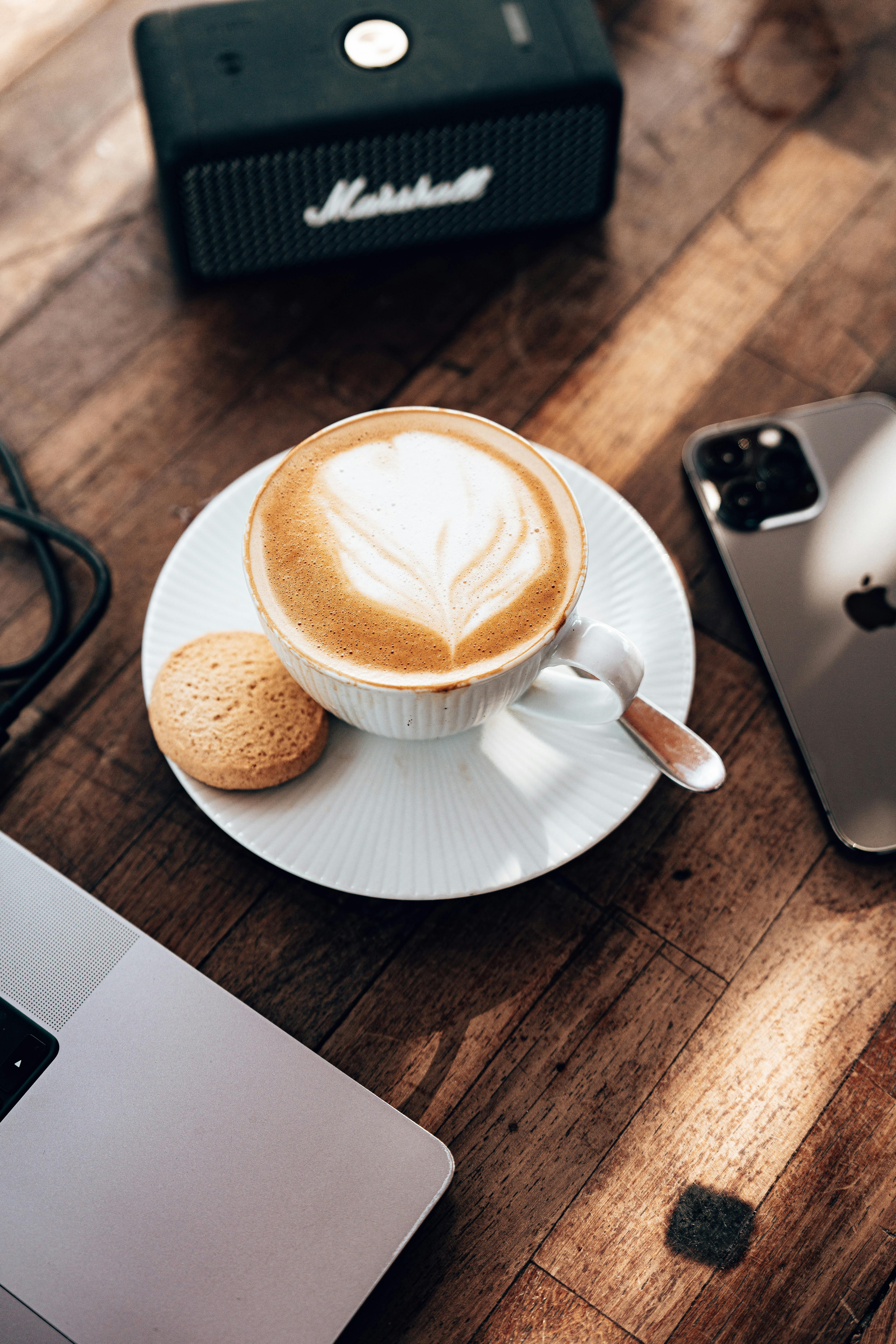High angle view of cappuccino and cookie next to smartphone and laptop on a wooden table.