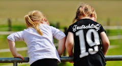 两个穿着休闲服的年轻女孩靠在篱笆上，探索着阳光明媚的田野