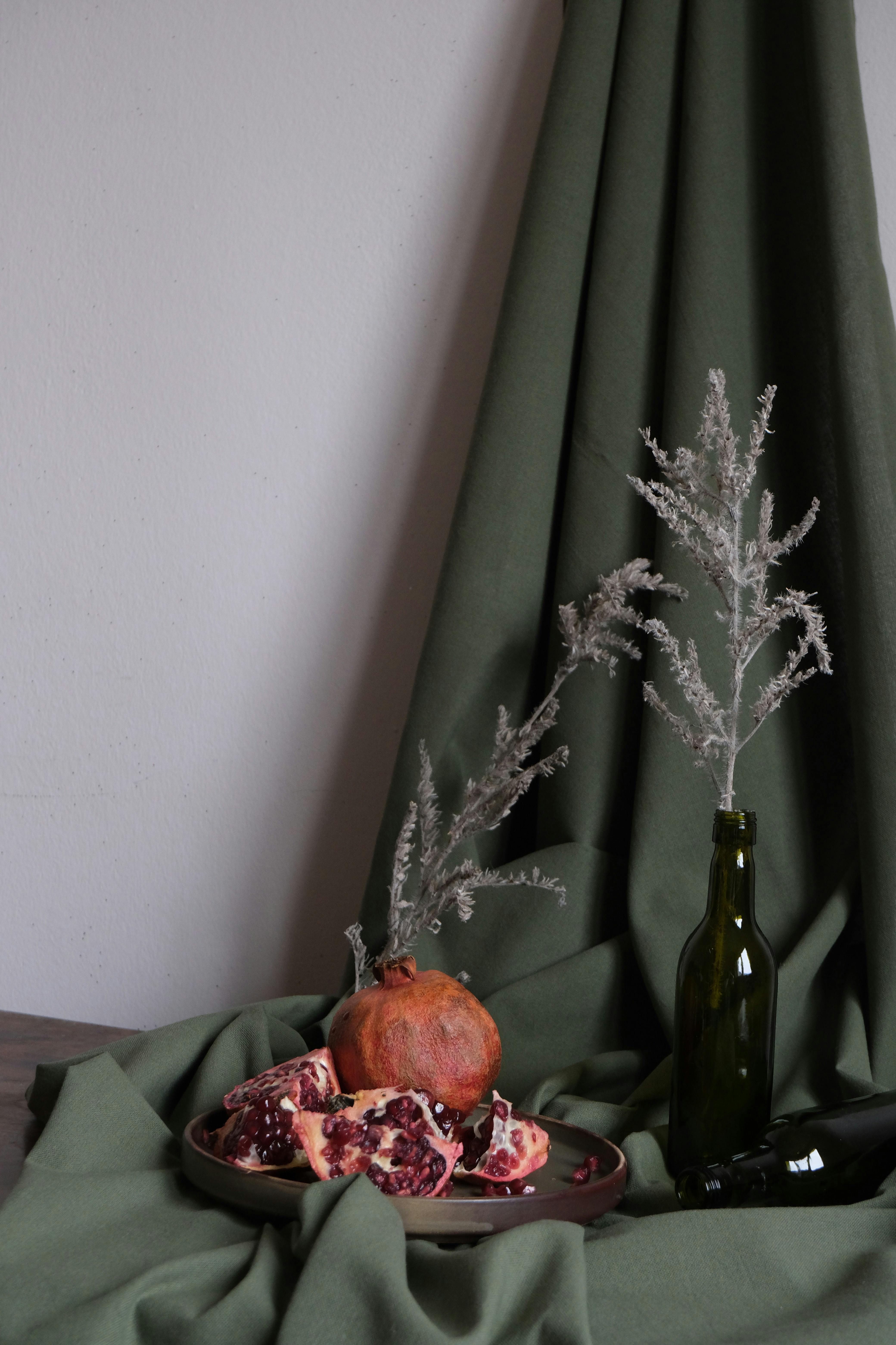 Artistic still life featuring a pomegranate, green drapery, and a glass bottle.