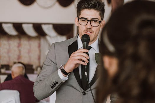 A man in a suit and glasses speaking into a microphone at a formal indoor event.