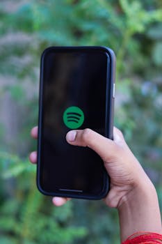 A person holding a smartphone displaying a music app in an outdoor setting with a green background.