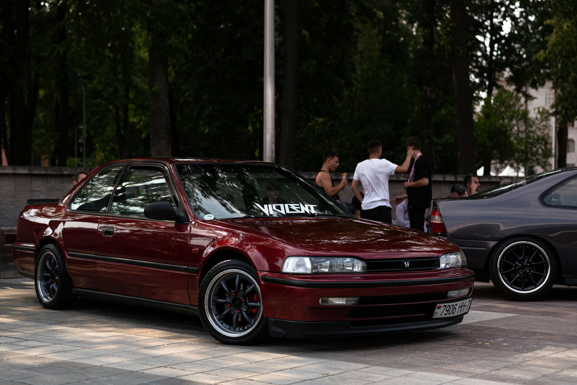 A red Honda Accord parked in an urban area with people standing nearby on a sunny day.